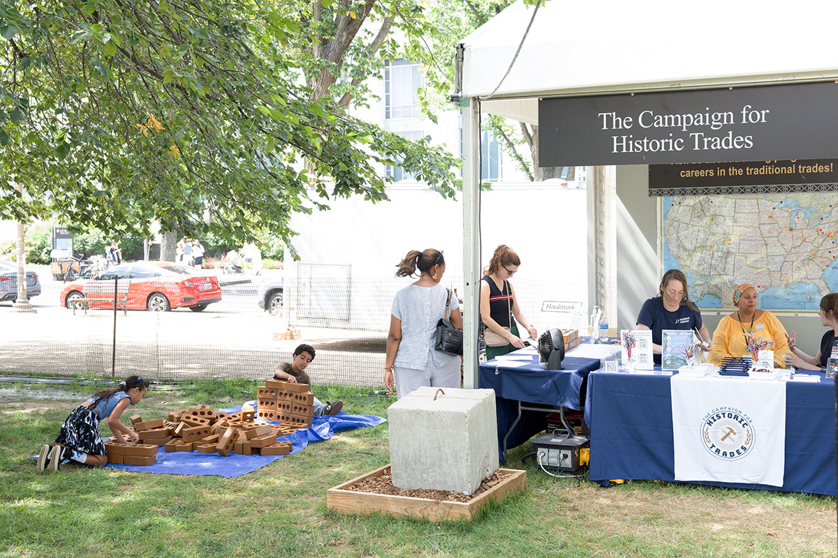 People sit under and stand around a tent with a banner across the top reading The Campaign for the Historic Trades. To the side, in the shade of a tree, two kids play with blocks on a blue tarp.