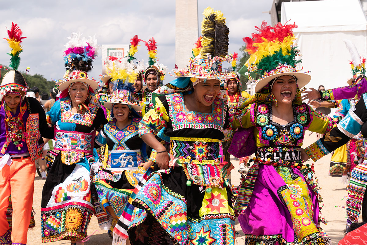 Several dancers in brightly embroidered regalia and tall feathered hats process down the National Mall, with the Washington Monument behind them.