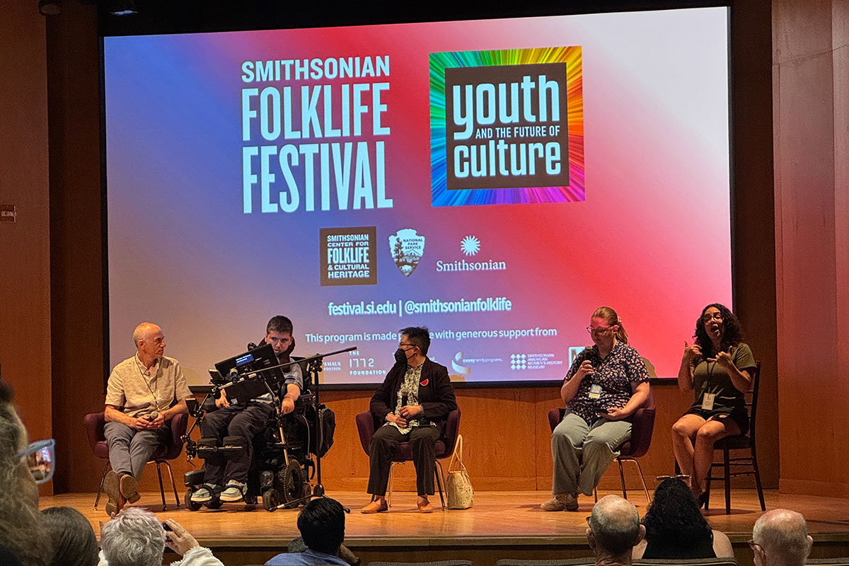Five people sit on stage in front of an audience. On one side, three people are in conversation. On the other, one person is audio describing and the other is interpreting the conversation into sign language.