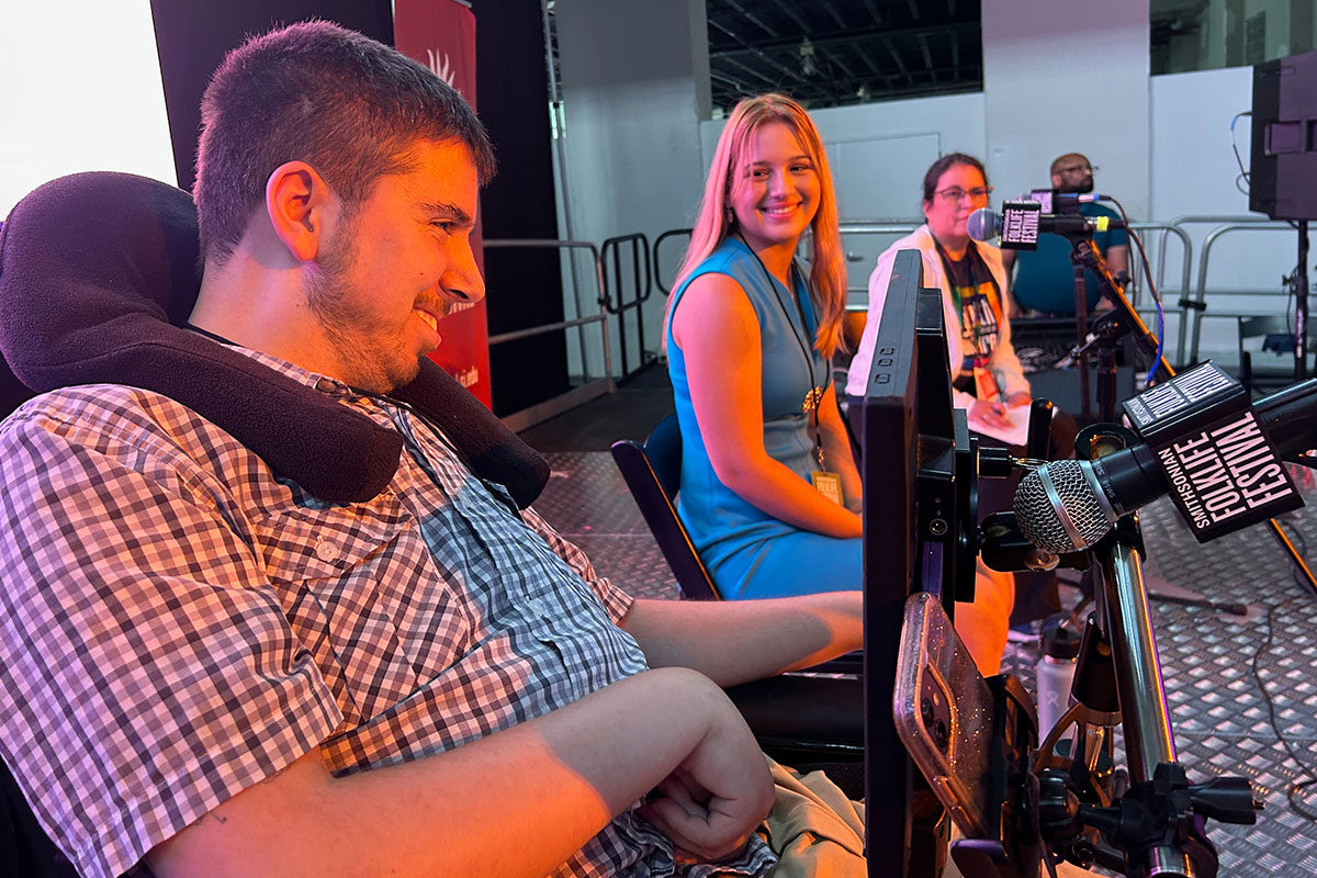 Three people sit on an illuminated stage. In the foreground, a young man in a plaid shirt smiles toward the audience. In front of him is a communication devide with a screen and a Folklife Festival microphone. The young woman next to him turns to look at him, also smiling. 