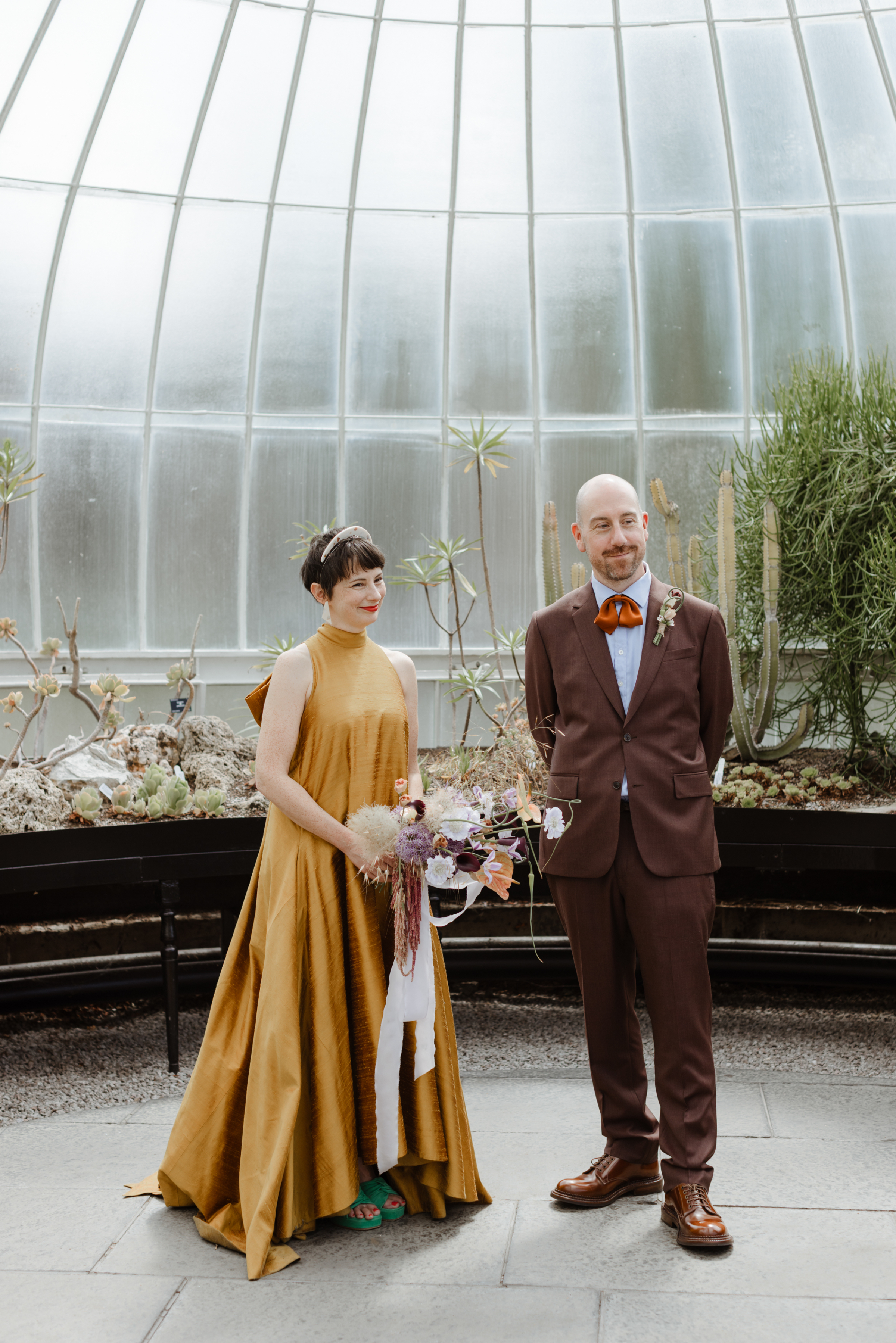 Bride in a non traditional mustard yellow dress with large bow at the back. She wears green platform open toe shoes and carries a bouquet she made herself. Groom in a brown suit and draped silk ribbon bow tie.