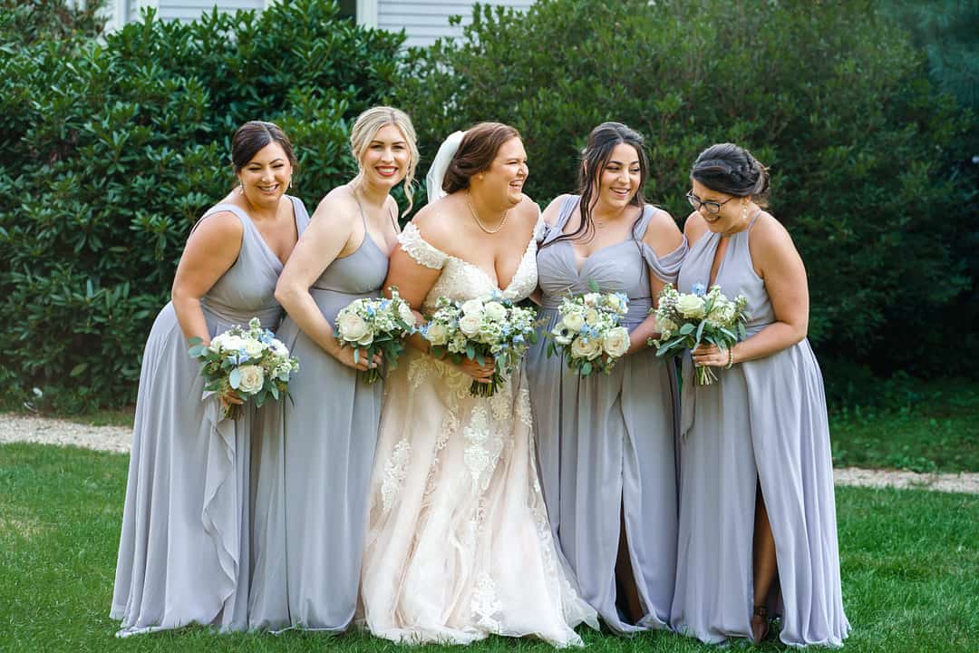 Plus-size bride in champagne off-shoulder lace fit-and-flare gown laughing with bridesmaids in light grey-blue dresses