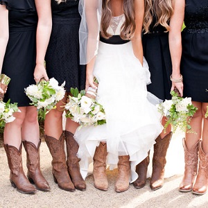 Close-up of bride and bridesmaids lifting skirts to show brown cowboy boots, bride in white ruffled gown and bridesmaids in black dresses, with white bouquets