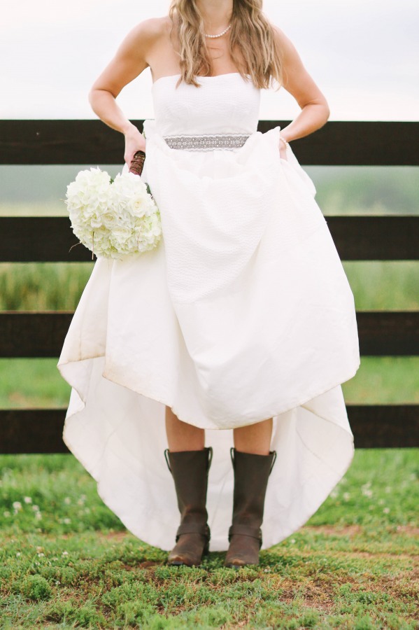 Bride in strapless a-line wedding gown with belted waist lifting skirt to reveal dark brown cowboy boots, holding white bouquet, at Lindsey Plantation South Carolina