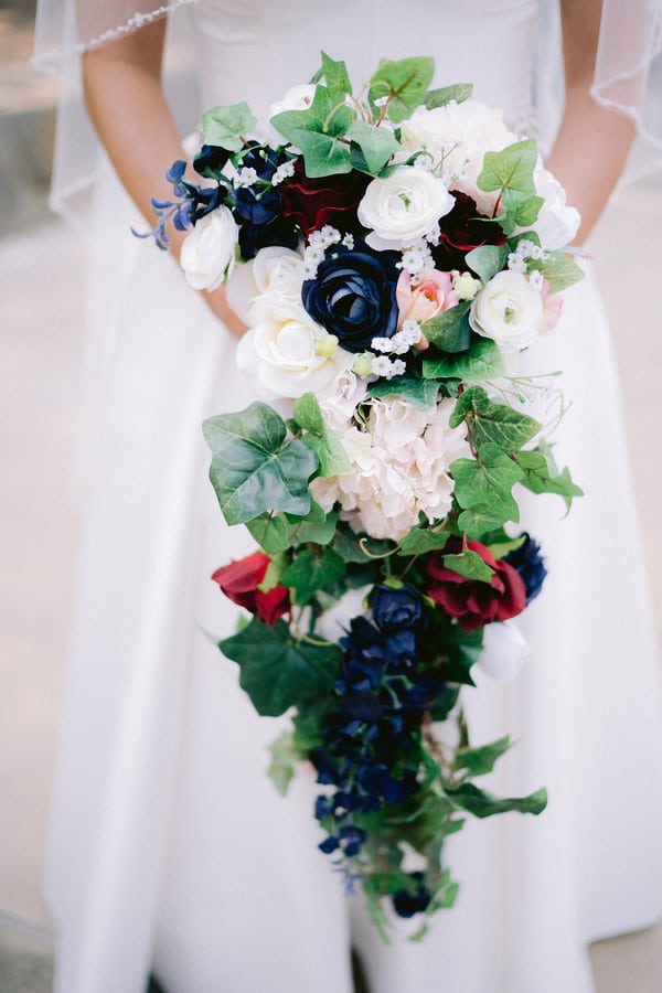 Bride holding a long dramatic cascade of white ranunculus, inky navy roses, blush hydrangea, and long trailing ivy against a white gown