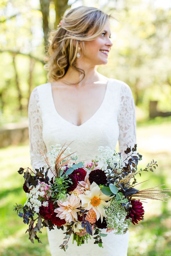 Bride in a long-sleeve lace gown outdoors holding an oversized bouquet of rich burgundy and peach dahlias with dark accent foliage, eucalyptus, and white filler blooms