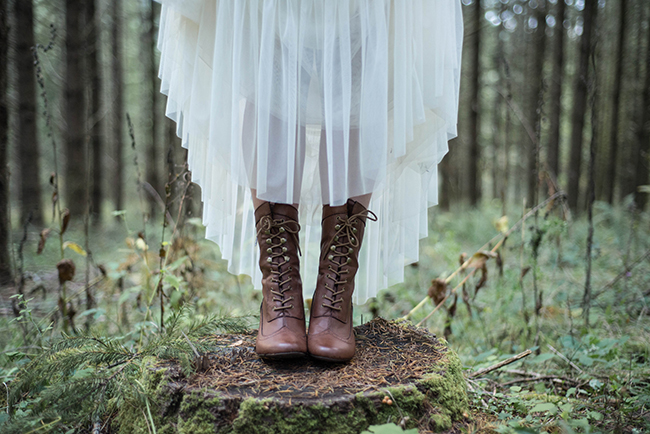 Bride in flowing white tulle wedding dress standing on tree stump in dark forest, wearing tall brown Victorian lace-up boots