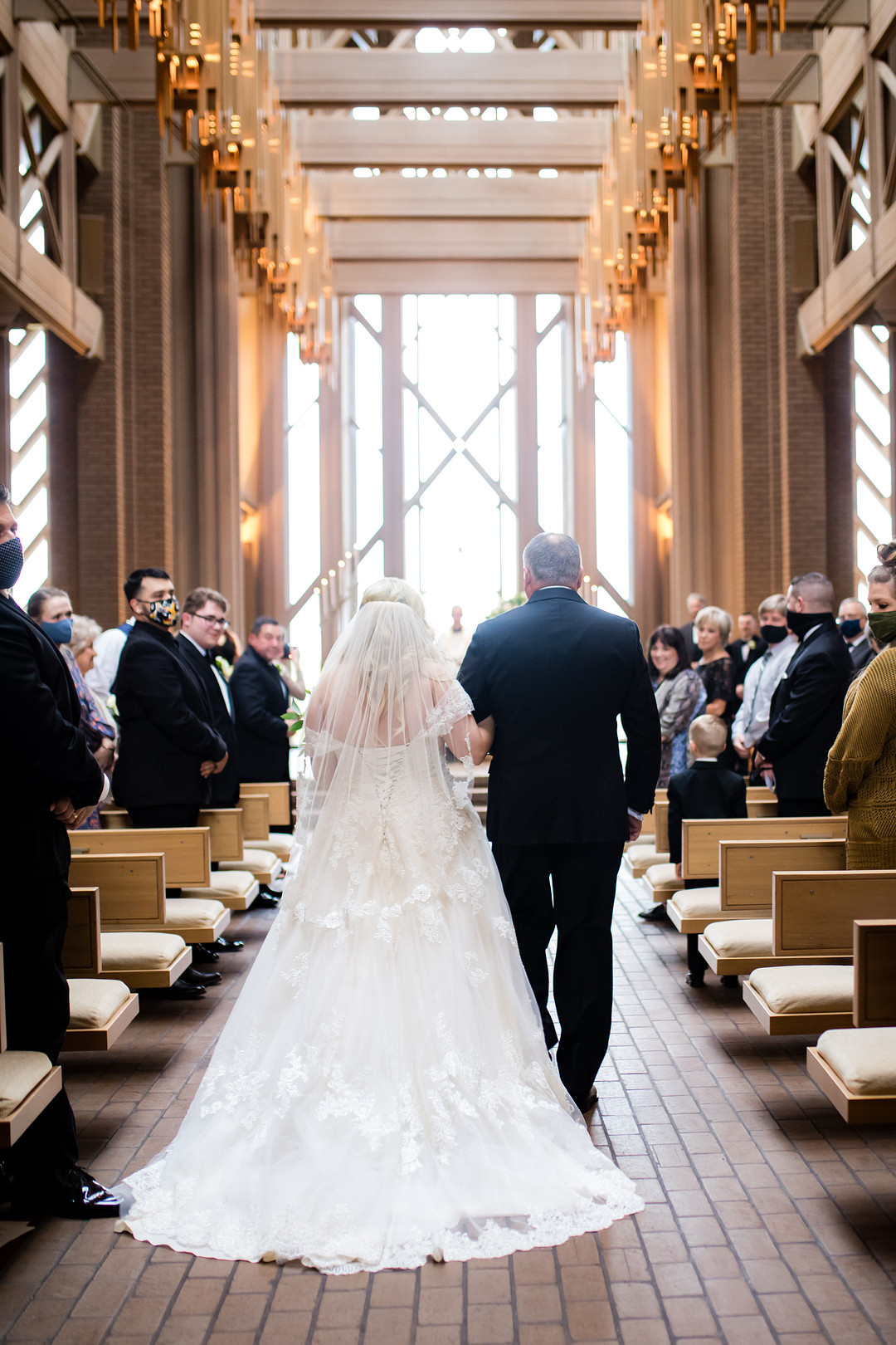 Bride in ivory off-shoulder lace wedding gown with chapel train walking down the aisle of a chapel in Fort Worth with groom and guests