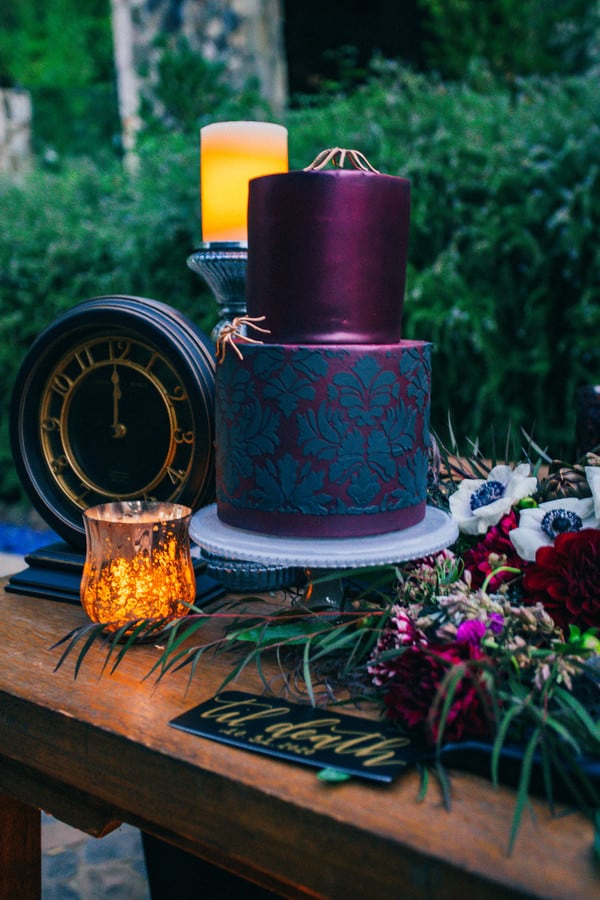 Two-tier navy blue fondant wedding cake with gold clockwork face detail, surrounded by dark burgundy anemones and greenery