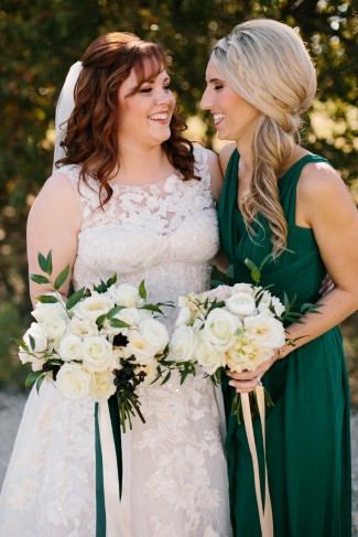 Plus-size bride in white lace A-line gown with scoop neckline and lace strap detail smiling with bridesmaid in forest green dress