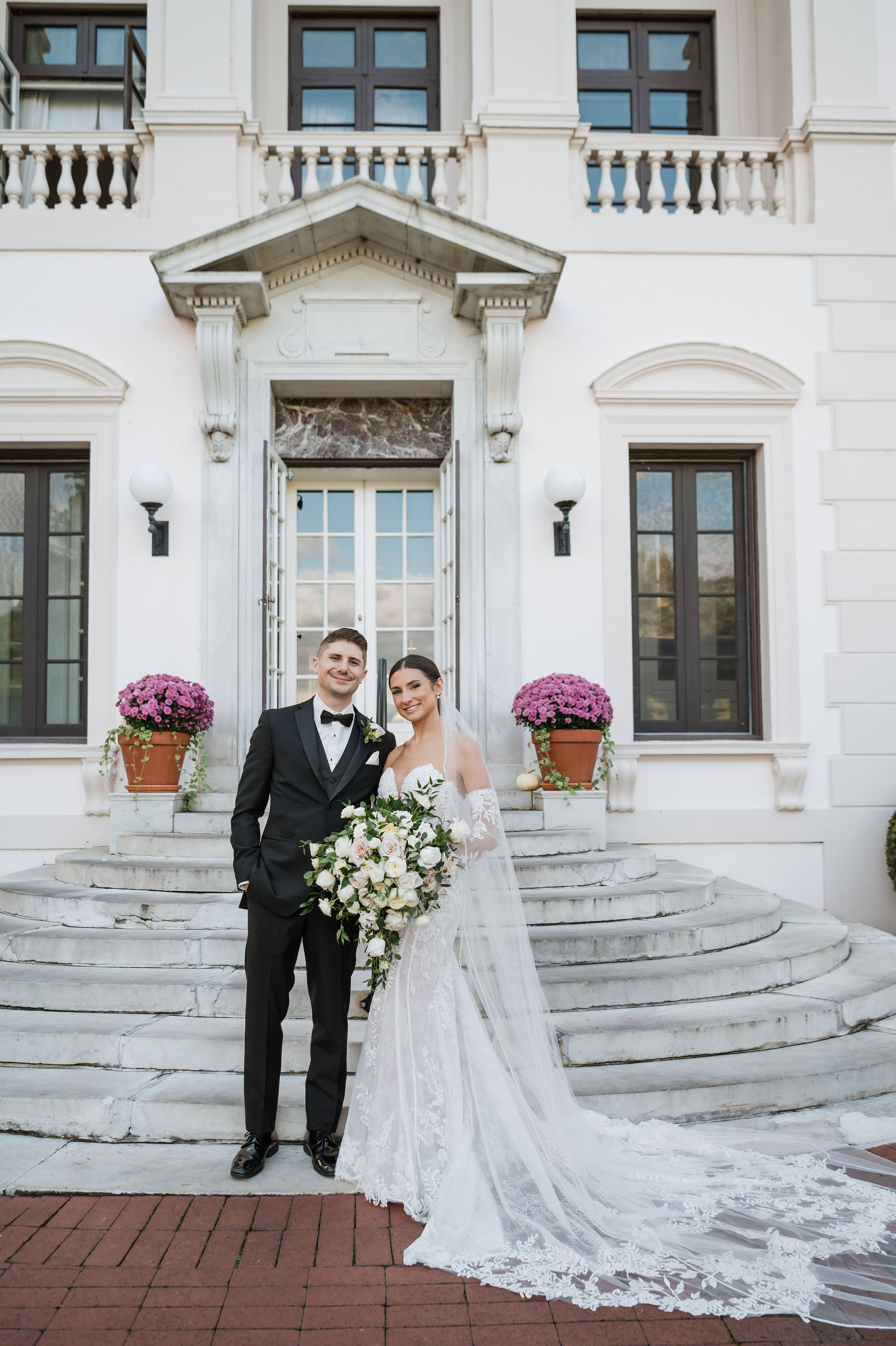 Couple standing on grand estate steps, bride in cathedral-length lace gown holding a large classic cascading bouquet of white and blush garden roses with trailing eucalyptus