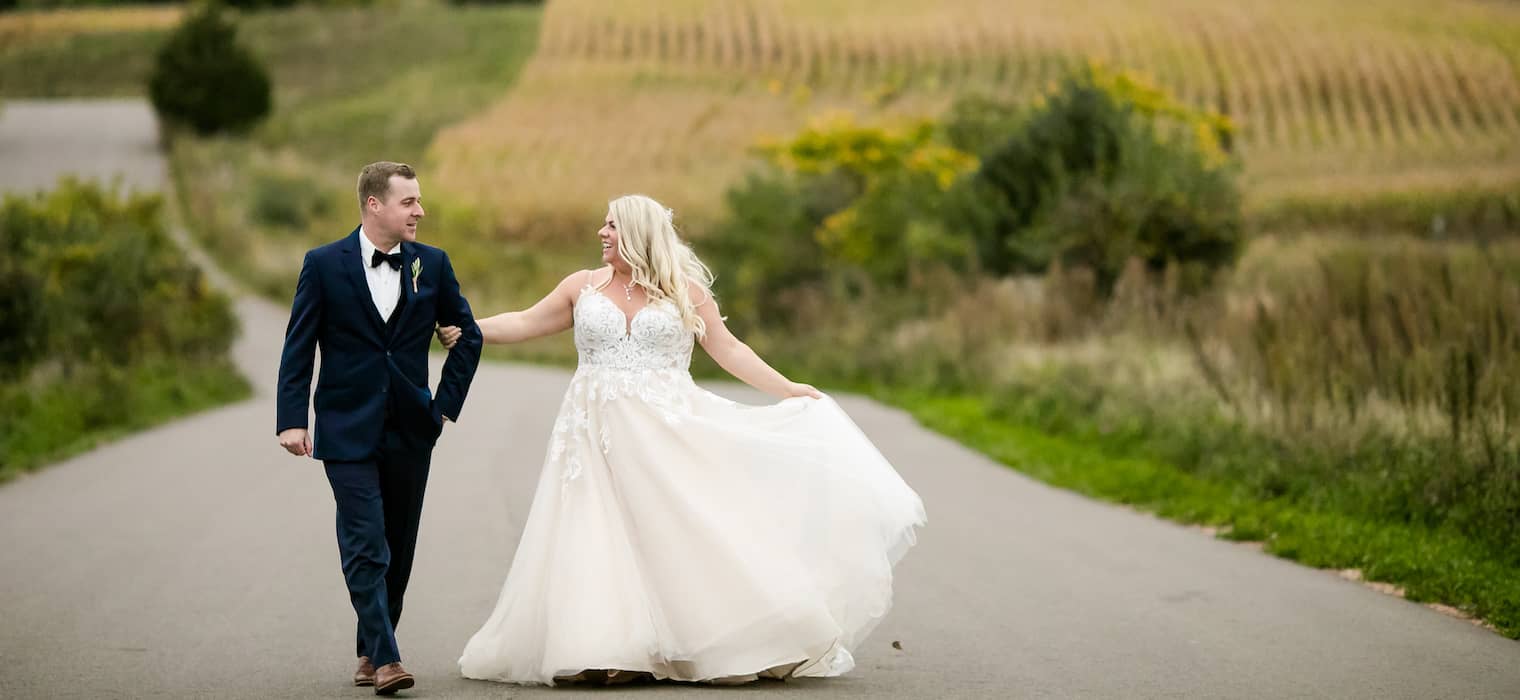 Bride in white ball gown with V-neckline, spaghetti straps, and lace bodice walking on a country road with groom