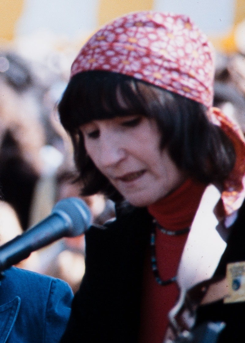 A woman with a brown bob and red handkerchief tied around her head plays acoustic guitar, singing into a mic outdoors.