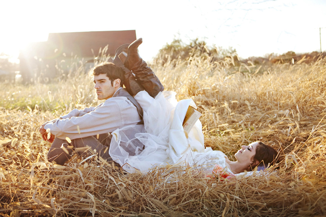 Bride in a-line lace and tulle wedding gown lying in golden wheat field with boots up, groom sitting beside her, at rustic anniversary session