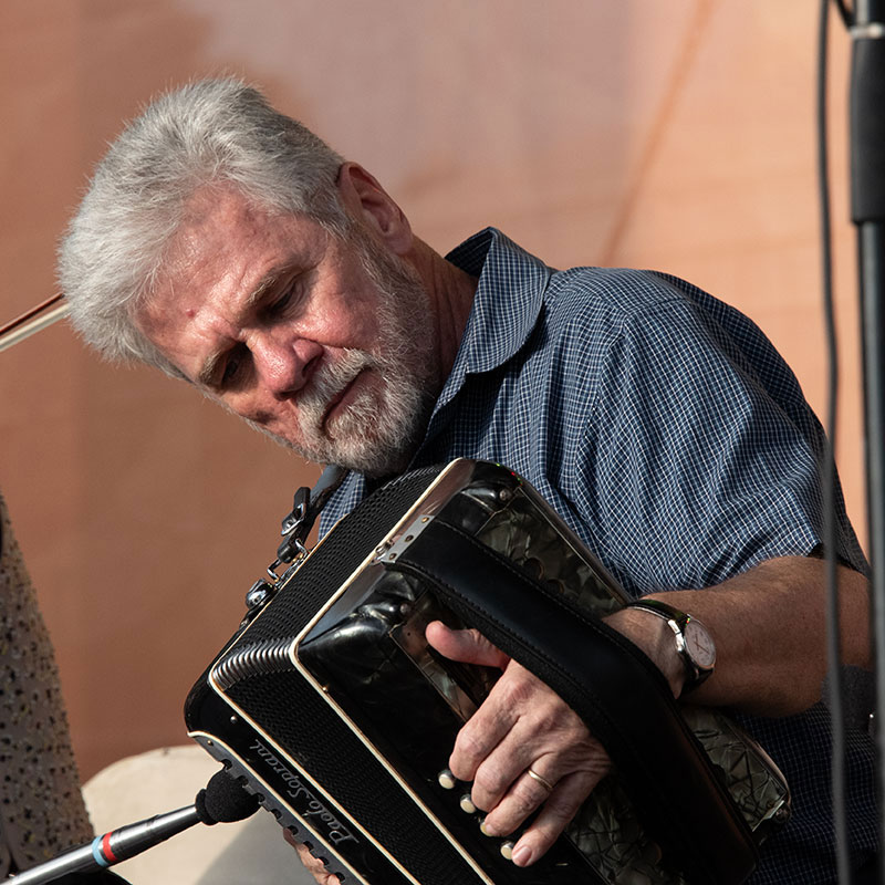 A man with short gray hair and beard playing a black accordion on stage, his head tilted toward the instrument.