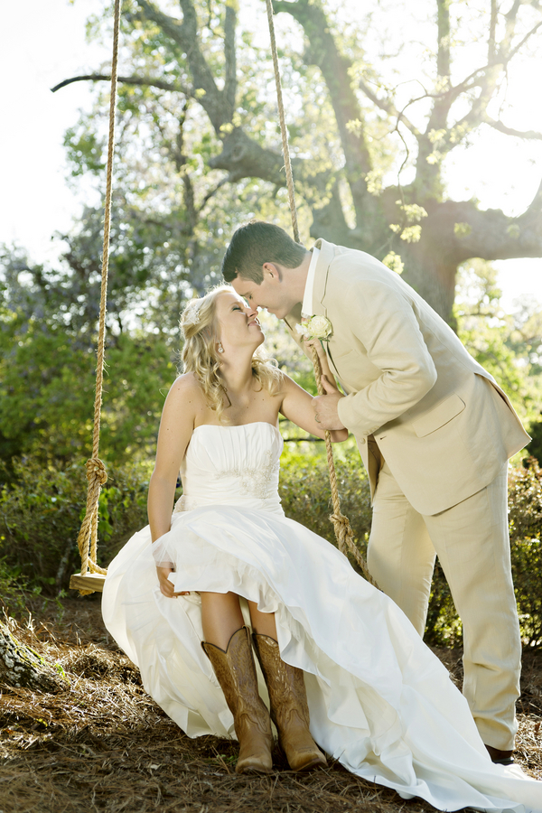 Bride in strapless sweetheart a-line wedding gown sitting on a swing with cowboy boots, groom leaning in to kiss her, at Fair Weather Farms Georgia