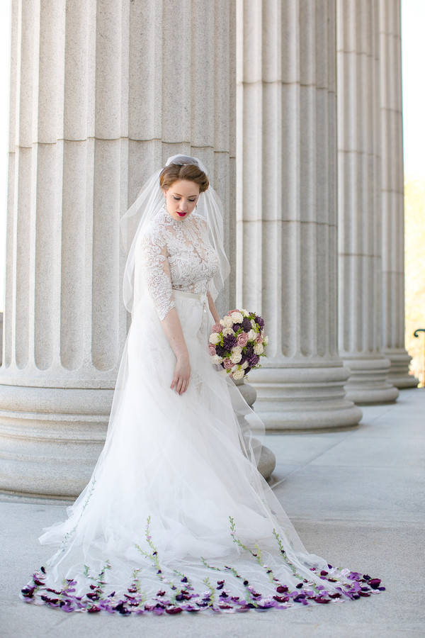 Bride in short-sleeve high-neck lace A-line gown beside stone columns at Minneapolis Institute of Art, with cathedral veil scattered with flower petals