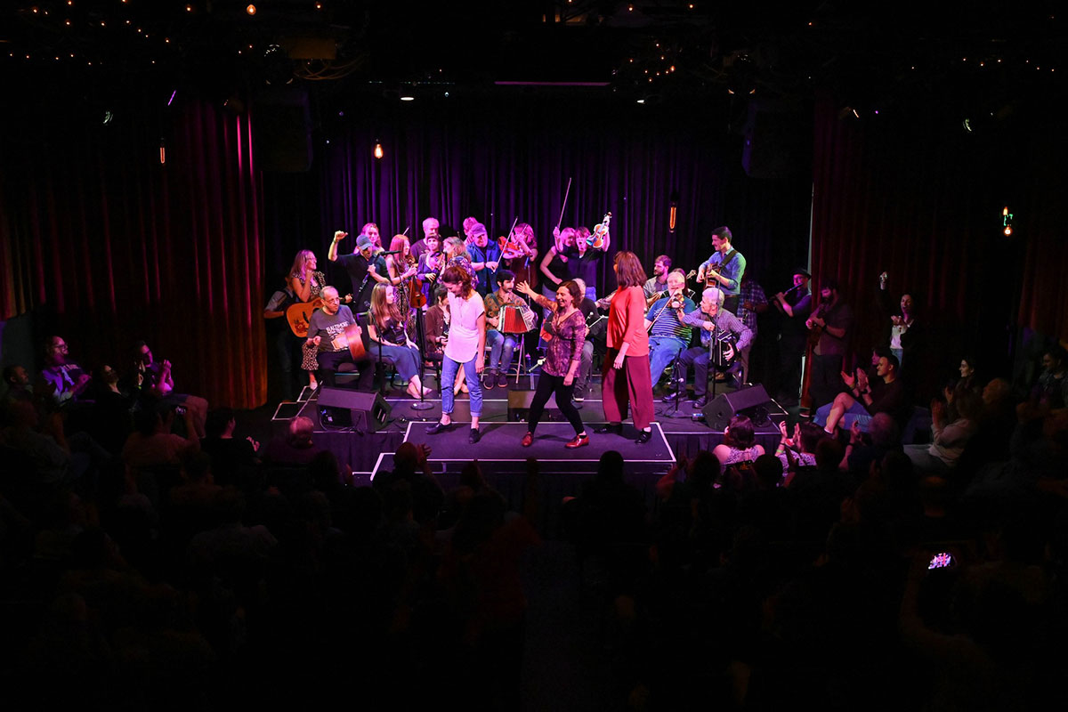 Musicians and dancers under purple lights on a small stage, surrounded by a crowd.