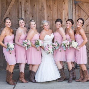 Bride in strapless sweetheart lace gown surrounded by bridesmaids in pink dresses with cowboy boots, in front of barn at Rivercrest Farm
