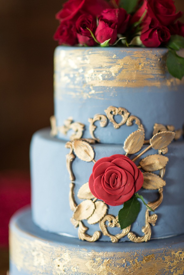 Three-tier dusty blue fondant wedding cake with intricate gold 3D embossed scrollwork, leaves, and a large red rose accent