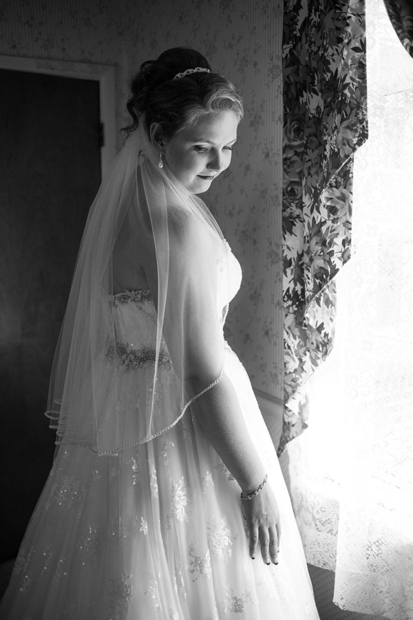 Bride in strapless white ball gown with beaded waistband and lace bodice photographed in black and white near a window with a cathedral veil