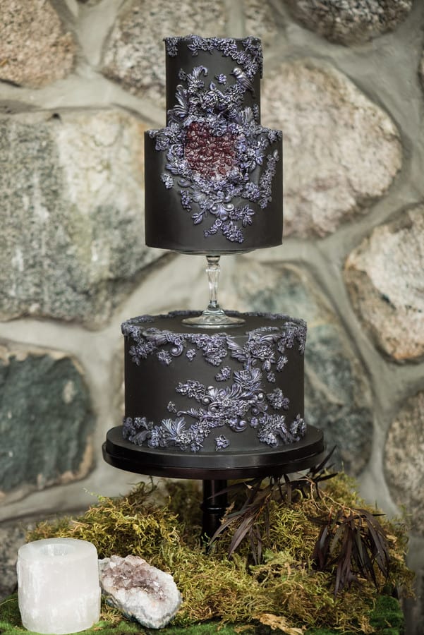 Two-tier black fondant wedding cake with intricate silver botanical lace embossing on a stone background