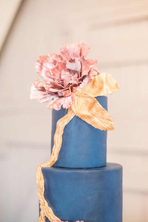 Two-tier navy blue fondant wedding cake with a large sculptured dusty pink sugar peony and gold leaf ribbon draped across the tiers