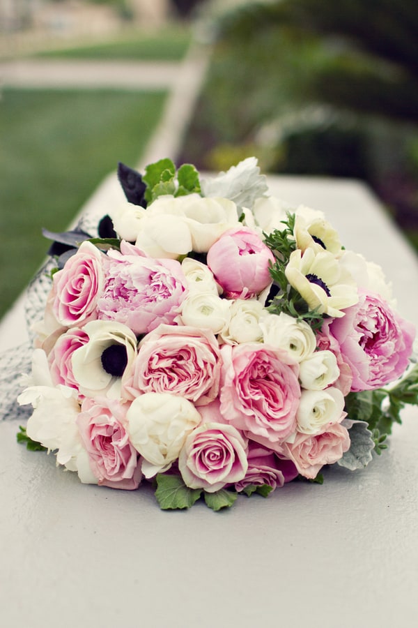 Lush bridal bouquet resting on a surface, featuring open pink peonies, white anemones with dramatic black centers, blush garden roses, ranunculus, and silver-green dusty miller