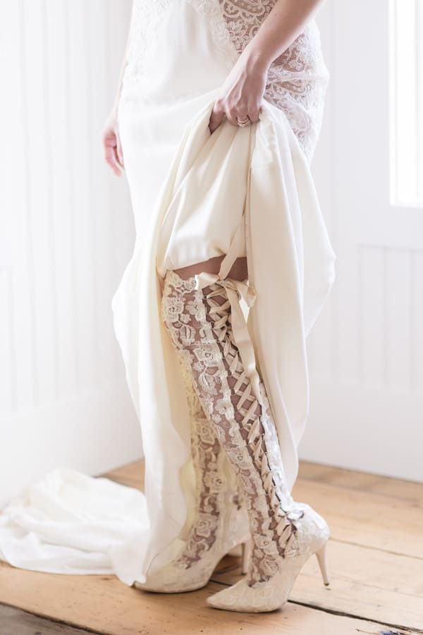 Bride in ivory lace sheath gown lifting skirt to show knee-high lace corset-tie boots by House of Elliot against bright window light