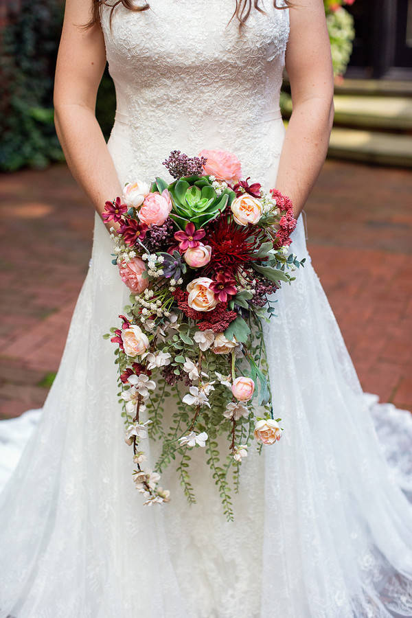 Bride holding a dramatic autumn cascade bouquet with blush garden roses, deep burgundy dahlias, succulents, and trailing waxflower against a white lace gown