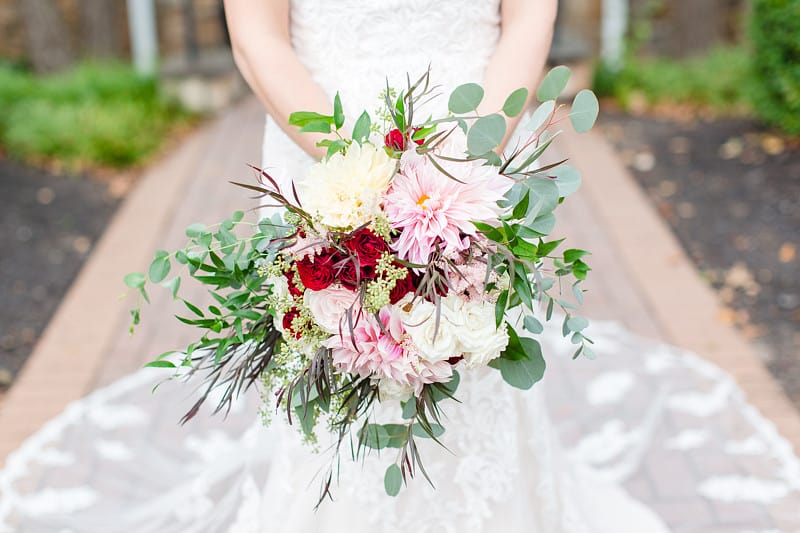 Bride at an outdoor country club wedding holding a full bouquet of blush dinner-plate dahlias, deep red garden roses, and dark accent foliage with trailing eucalyptus
