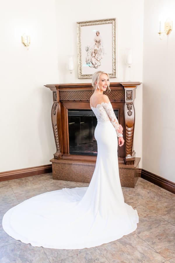 Bride in off-shoulder long-sleeve crepe and lace mermaid gown standing by ornate fireplace