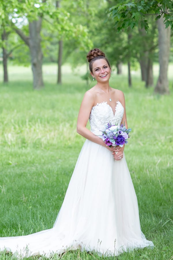 Smiling bride in strapless beaded lace A-line wedding gown holding bouquet outdoors in green field