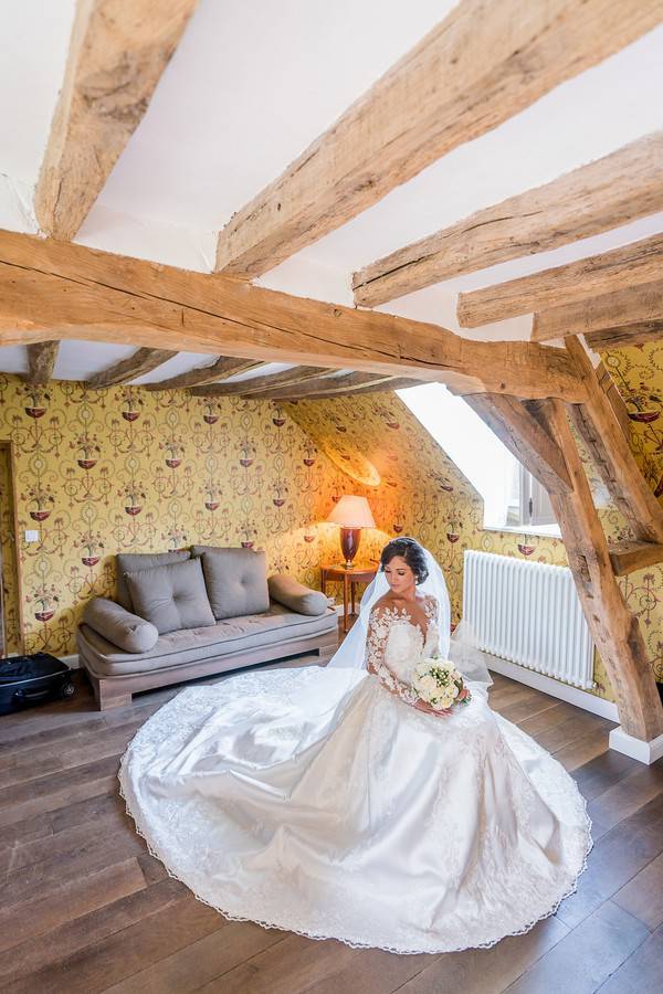 Bride in long-sleeve illusion lace ballgown with cathedral train sitting in French chateau room with exposed wooden beams