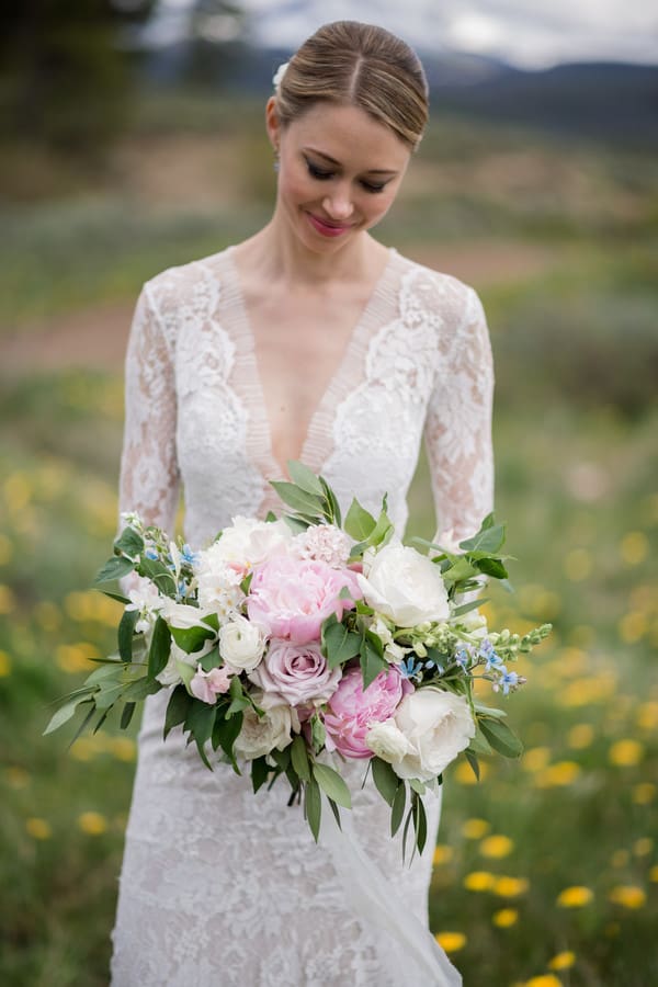 Bride in deep-V lace gown standing in a wildflower meadow with mountains behind, holding a full generous bouquet of blush peonies, mauve garden roses, and soft blue forget-me-nots