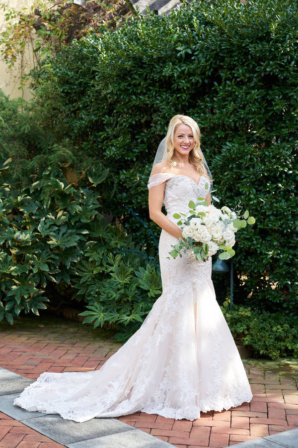 Bride in off-shoulder ivory lace mermaid gown standing in front of lush greenery at botanical garden wedding