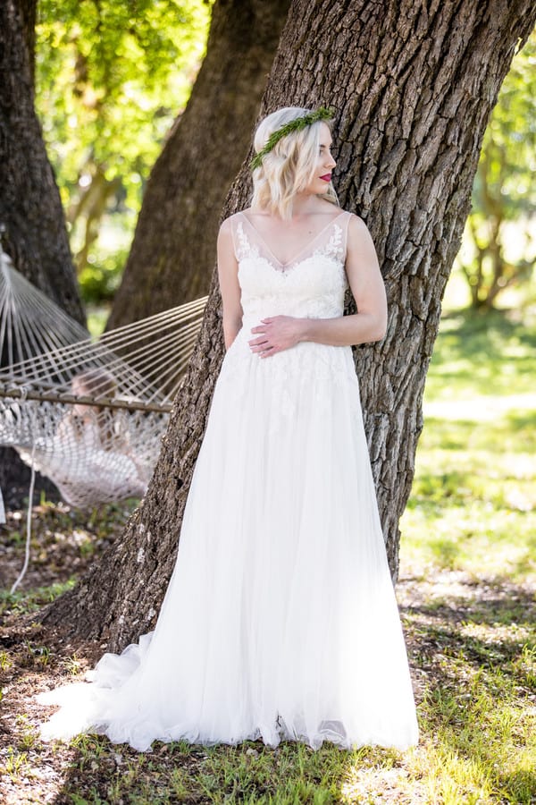 Bride in illusion lace A-line gown with thin straps and greenery crown leaning against a large tree trunk in an outdoor Texas garden, hammock visible in the background
