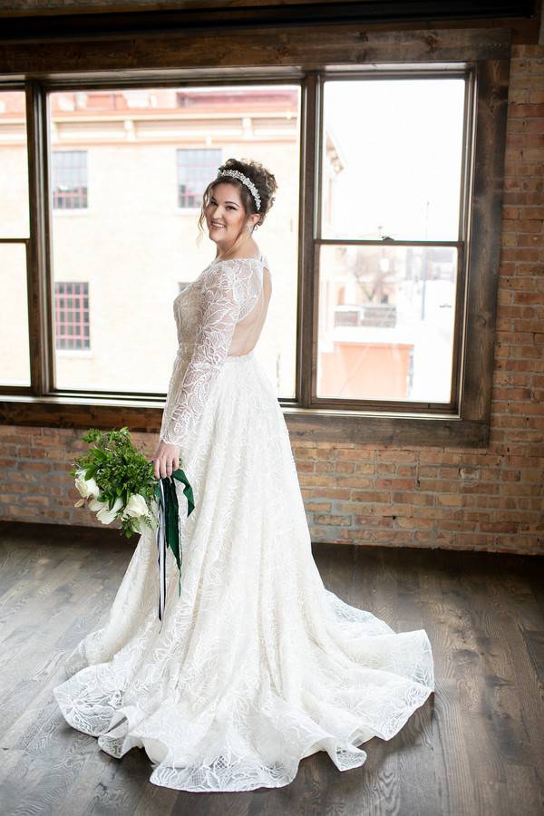 Bride in long-sleeve boat-neck lace A-line gown with open back holding white bouquet, standing by industrial loft windows in Woodstock, Illinois