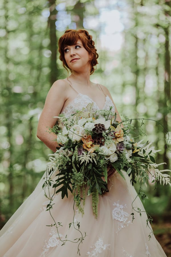 Bride in a blush ball gown in a forest setting holding an enormous cascading bouquet with succulents, orchids, ferns, and trailing vines overflowing over both arms