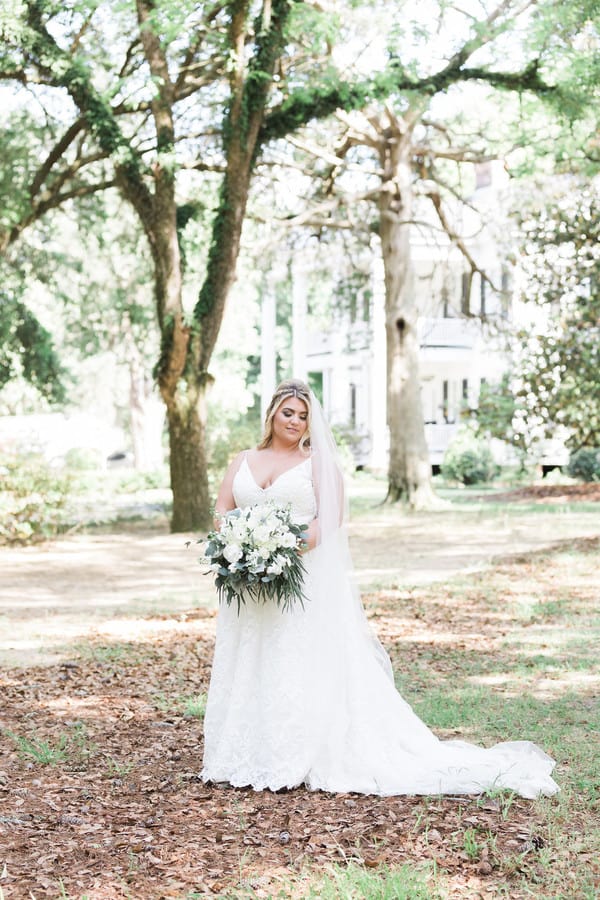 Plus-size bride in white lace A-line gown with spaghetti straps and V-neck, standing outdoors with cathedral veil flowing behind her