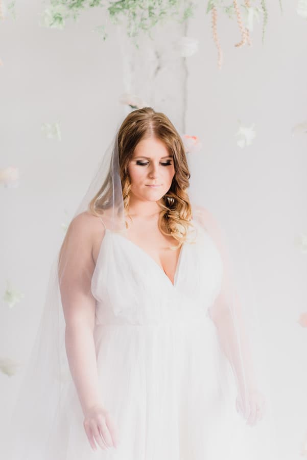 Plus-size bride in minimalist white V-neck slip gown with spaghetti straps and cathedral veil in a bright floral studio setting