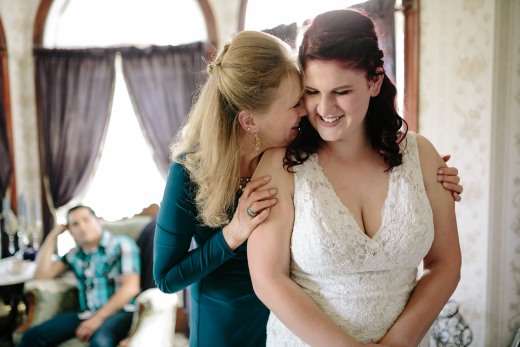 Plus-size bride in white lace V-neck wedding gown being embraced by mother during getting-ready moment in a California wedding venue