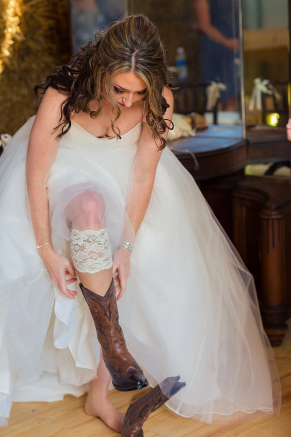 Bride in white ballgown getting ready, lifting skirt to show brown cowboy boot with lace garter, at Carlee Farm North Carolina wedding