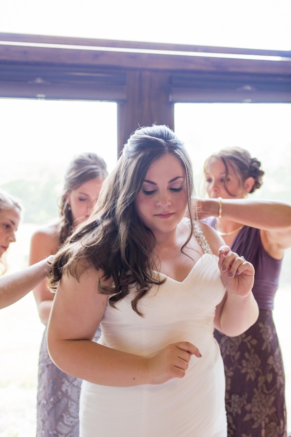 Plus-size bride in white wedding gown with thin straps being helped by bridesmaids during getting-ready at a backyard Midwest wedding