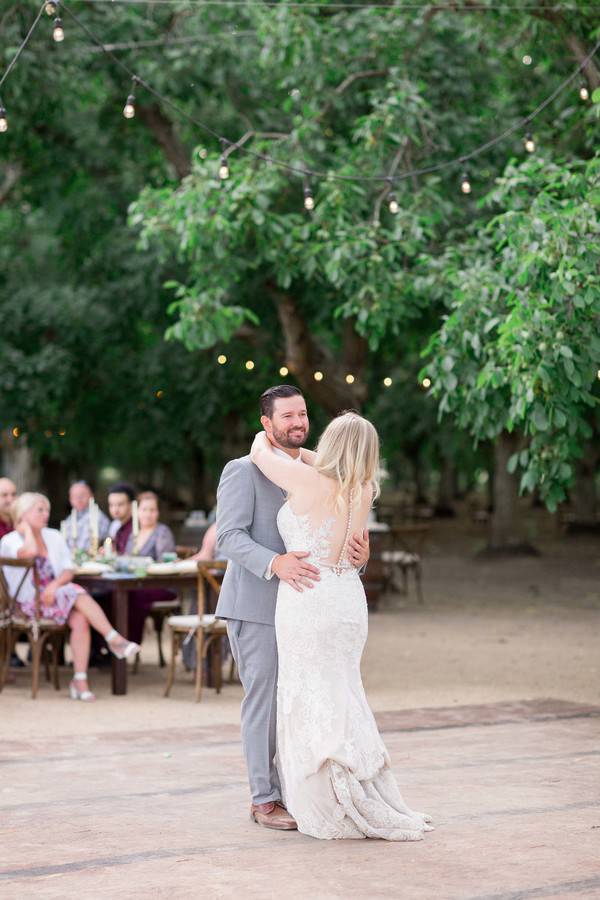 Bride in white lace fit-and-flare gown with open back and spaghetti straps dancing first dance with groom at an outdoor wedding under string lights