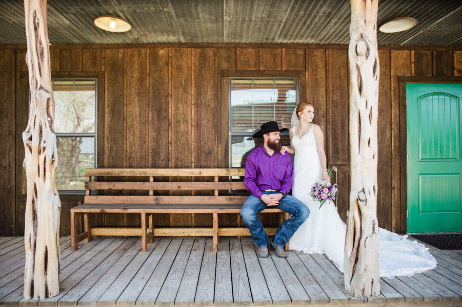 Bride in white wedding gown and groom in cowboy hat, purple western shirt and jeans, sitting on ranch porch at Katzfey Ranch South Texas wedding