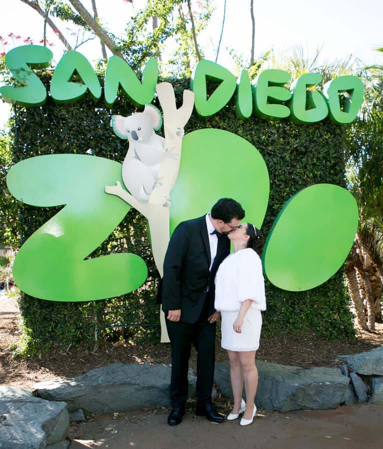 Bride in short white dress with white fur stole kissing groom in front of the San Diego Zoo sign