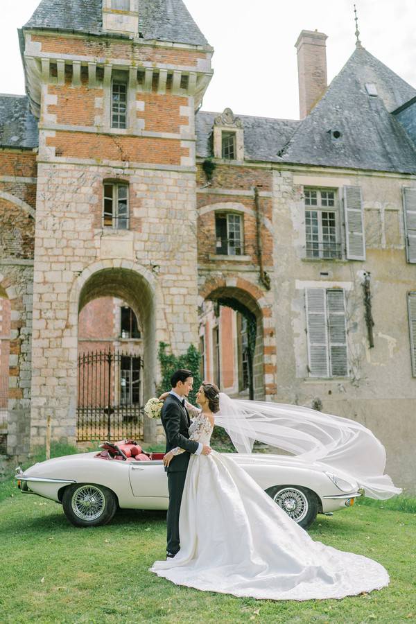Bride and groom posing outside French chateau next to vintage car, bride in white long-sleeve v-neck ballgown with veil caught in wind