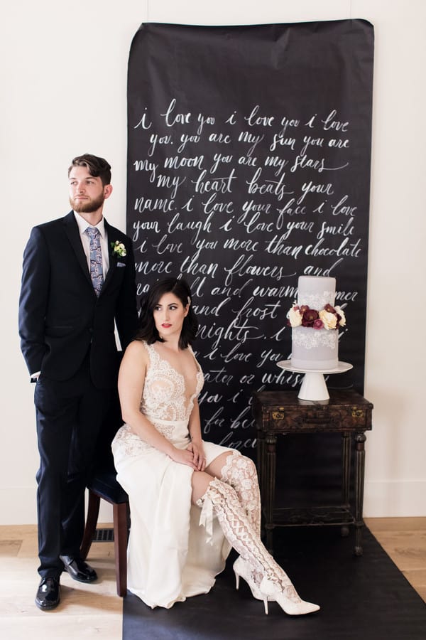 Bride in lace sheath gown with knee-high lace boots seated beside groom in dark suit against large handwritten chalkboard backdrop at distillery wedding shoot