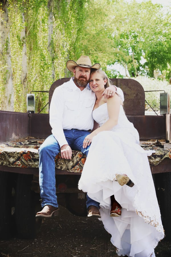 Bride in white high-low wedding gown and groom in cowboy hat sitting together on the tailgate of an old pickup truck at Indian Springs Ranch Nevada wedding